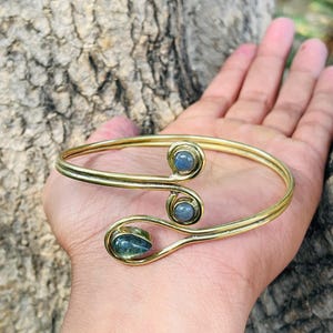 Pode incluir: Uma pulseira de metal dourado com três pedras preciosas azul-esverdeadas. A pulseira tem um design curvo com as pedras preciosas em engastes circulares. A pulseira é mostrada na mão de uma pessoa.