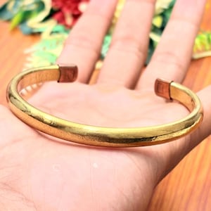 May include: A gold-toned metal cuff bracelet with a smooth, polished finish and a rounded profile. The bracelet features an open design with two distinct copper-colored rectangular end caps at each terminal point of the circular band.
