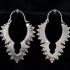 May include: A pair of silver-toned earrings. Each earring features a curved, open design with a series of small, round embellishments along the edges. The earrings have a hook closure and a decorative, openwork design.