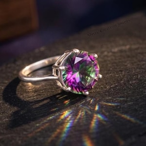 Puede incluir: Un anillo de plata con una gran piedra preciosa redonda y multicolor. La piedra preciosa muestra tonos de morado, verde y azul. El anillo está sobre una superficie oscura, con la luz reflejada en la piedra.