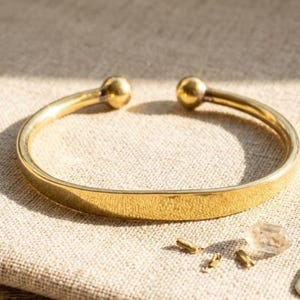 May include: A gold-coloured cuff bracelet with rounded ball ends. The bracelet is open at the bottom and rests on a neutral-coloured surface. Small gold-coloured jewellery findings and a clear crystal are also present.