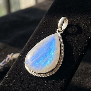 May include: A teardrop-shaped pendant with a shimmering, iridescent moonstone. The stone is set in a silver-colored metal with a twisted rope design around the edge. The pendant has a small loop at the top for a chain.
