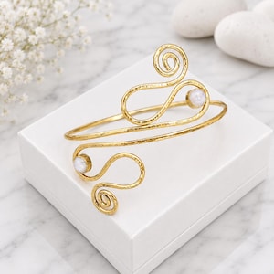 May include: A gold-toned metal cuff bracelet featuring a hammered texture and an open-ended design. The ends of the bracelet are shaped into decorative swirls, with each terminal point set with a small, round, iridescent white cabochon stone.