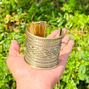 May include: A wide gold-toned metal cuff bracelet featuring a central braided texture flanked by horizontal bands of intricate geometric patterns. The bracelet has an open back design and a polished finish, showcasing detailed metalwork throughout the piece.