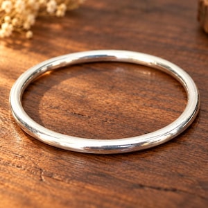 May include: A polished silver bangle bracelet with a smooth, rounded surface and a continuous circular shape. The metal has a reflective finish that catches the light, creating bright highlights along the top edge of the solid, seamless band.