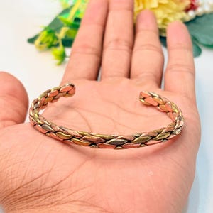 May include: A cuff bracelet featuring a braided design made of alternating strands of copper, brass, and silver-toned metal. The open-ended bangle has a textured, woven appearance with a polished finish, showcasing a mix of warm and cool metallic hues.