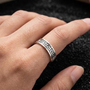 Puede incluir: Un anillo plateado con un patrón de greca. El anillo se lleva en un dedo, mostrando el intrincado diseño. La superficie del anillo refleja la luz, resaltando los detalles del patrón geométrico. El anillo es una pieza de joyería.