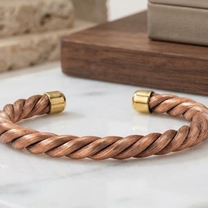 May include: A copper-colored cuff bracelet featuring a twisted wire design. The bracelet has a metallic finish and is capped at both open ends with smooth, polished gold-toned cylindrical metal accents, creating a continuous circular shape.