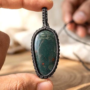 May include: A dark green gemstone pendant with red flecks, encased in a black macrame design. The pendant is oval-shaped and held between fingers, showcasing the intricate knotwork and natural stone. The overall theme is handcrafted jewelry.