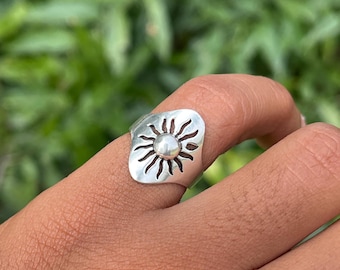 May include: A silver ring featuring a sun design. The ring has a rectangular face with a cut-out sunburst pattern surrounding a central, raised circular element. The ring is worn on a finger, with a blurred green background.