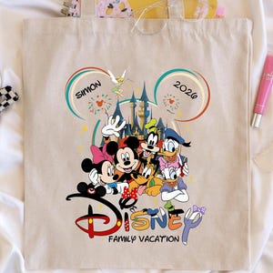 K&ouml;nnte beinhalten: Beige Tragetasche mit einem Disney-Familienurlaubsdesign. Das Design umfasst Micky und Minnie Maus, Goofy, Donald Duck und andere Charaktere. Der Text "SIMON 2026" und "Disney Family Vacation" sind ebenfalls sichtbar.
