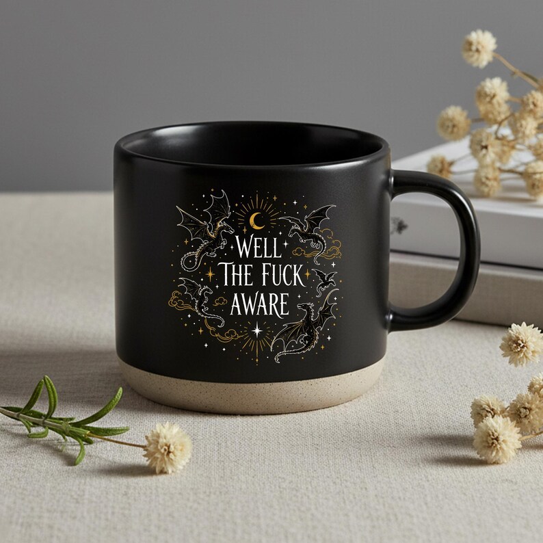 May include: Black ceramic mug with a beige base. The mug features the text "WELL THE FUCK AWARE" in white and gold, surrounded by dragon illustrations. The mug has a curved handle and is set against a neutral background.