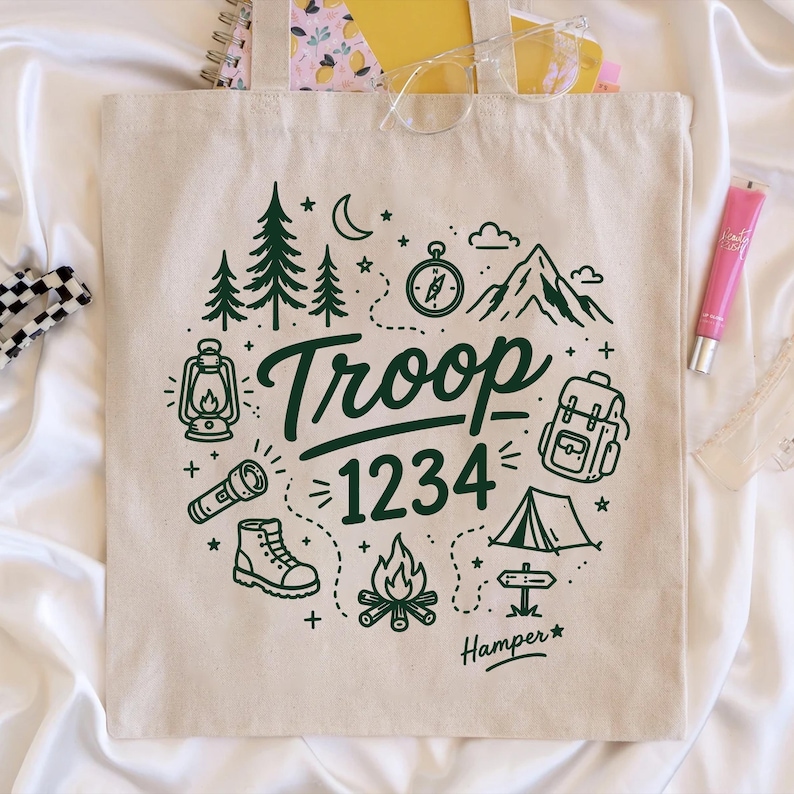 Op de afbeelding: Beige canvas draagtas met een donkergroen kampeermotief. Het ontwerp omvat bomen, een kompas, bergen, een rugzak, een tent en de tekst "Troop 1234". Het woord "Hamper" is ook opgenomen.