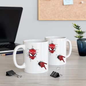 Puede incluir: Dos tazas de cer&aacute;mica blanca con un dise&ntilde;o de Spider-Man rojo y negro. El dise&ntilde;o incluye la m&aacute;scara de Spider-Man y una mano, con un efecto de pared agrietada. Las tazas est&aacute;n sobre una superficie clara con material de oficina y un port&aacute;til en el fondo.