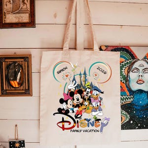 K&ouml;nnte beinhalten: Beige Canvas-Tragetasche mit farbenfrohem Disney-Design. Das Design zeigt Micky und Minnie Maus, Donald und Daisy Duck, Goofy und Pluto. Der Text lautet "Disney Family Vacation" mit dem Namen "Simon" und dem Jahr "2026".