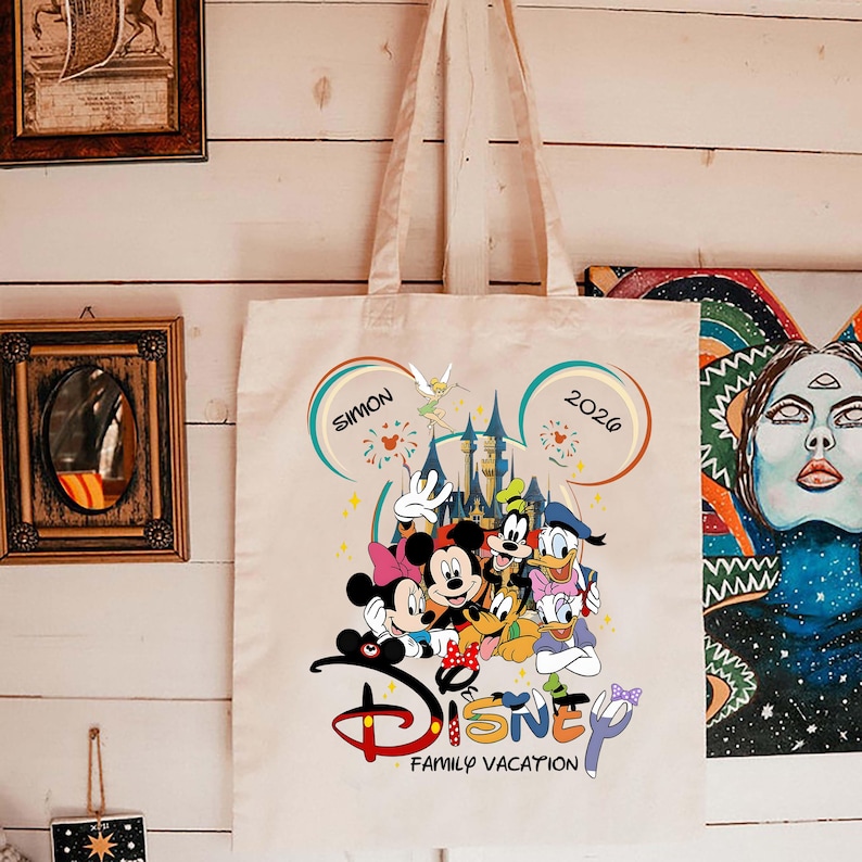 K&ouml;nnte beinhalten: Beige Canvas-Tragetasche mit farbenfrohem Disney-Design. Das Design zeigt Micky und Minnie Maus, Donald und Daisy Duck, Goofy und Pluto. Der Text lautet "Disney Family Vacation" mit dem Namen "Simon" und dem Jahr "2026".