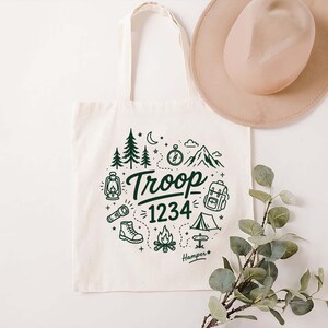 Op de afbeelding: Cr&egrave;mekleurige canvas draagtas met een groen kampeerthema. Het ontwerp bevat bomen, bergen, een kompas en de tekst "Troop 1234". Een beige hoed hangt boven de tas, en een takje groen staat op de voorgrond.