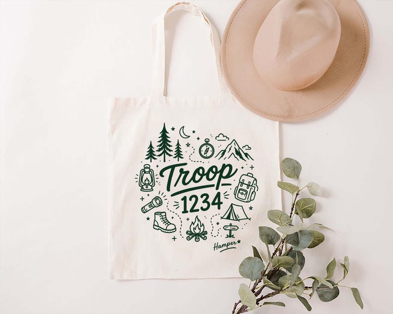 Op de afbeelding: Cr&egrave;mekleurige canvas draagtas met een groen kampeerthema. Het ontwerp bevat bomen, bergen, een kompas en de tekst "Troop 1234". Een beige hoed hangt boven de tas, en een takje groen staat op de voorgrond.