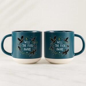 May include: Two teal ceramic mugs with white bases. Each mug features a design with black dragons, a crescent moon, stars, and the text "WELL THE FUCK AWARE". The mugs have a rounded shape and a large handle.