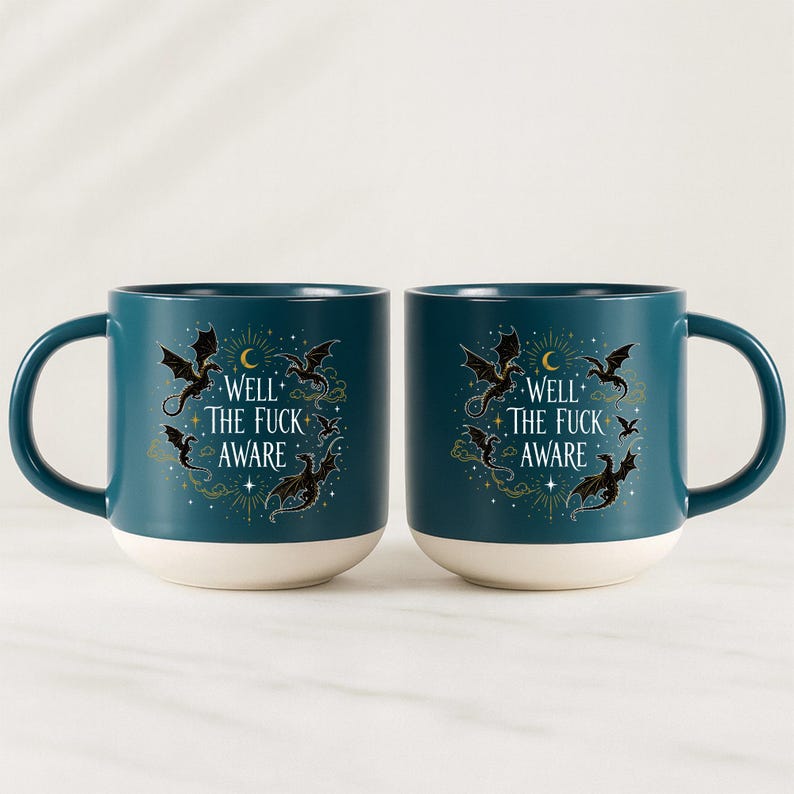 May include: Two teal ceramic mugs with white bases. Each mug features a design with black dragons, a crescent moon, stars, and the text "WELL THE FUCK AWARE". The mugs have a rounded shape and a large handle.