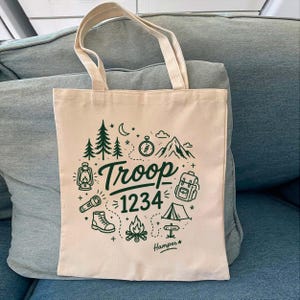 Op de afbeelding: Cr&egrave;mekleurige canvas draagtas met groene kampeerafbeeldingen, waaronder bomen, een kompas en een tent. De tekst "Troop 1234" is prominent aanwezig. De tas heeft twee handvatten.