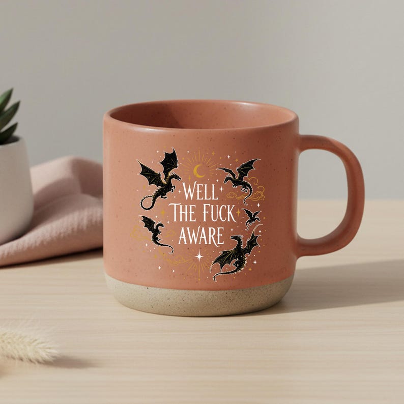 May include: A terracotta-coloured ceramic mug with a cream-coloured base. The mug features a design of black dragons, a crescent moon, and the text "WELL THE FUCK AWARE". The mug has a large handle for easy gripping.
