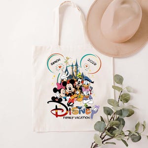 K&ouml;nnte beinhalten: Cremefarbene Tragetasche mit farbenfrohem Disney-Familienurlaubsdesign. Das Design umfasst Micky Maus, Minnie Maus, Donald Duck, Goofy und andere Charaktere. Der Text "Disney Family Vacation" befindet sich darunter.