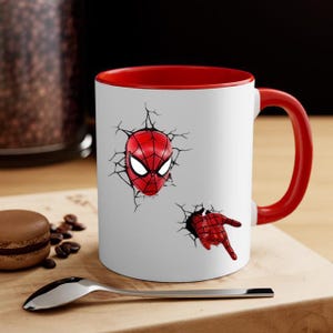 Puede incluir: Taza de cer&aacute;mica blanca con interior y asa rojos. La taza presenta un dise&ntilde;o de Spider-Man, con la m&aacute;scara y la mano del personaje aparentemente rompiendo la superficie. Una cuchara y un pastelito est&aacute;n en primer plano.