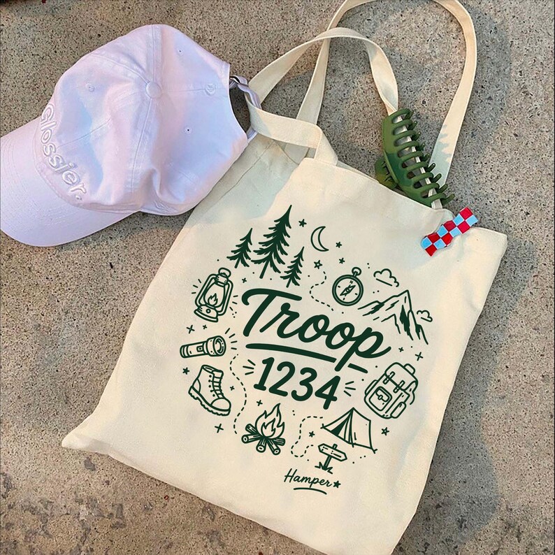 Op de afbeelding: Cr&egrave;me canvas draagtas met een groene campingafbeelding met de tekst "Troop 1234" en een witte baseballpet. De tas heeft een groene haarclip en een rood, wit en blauw haarspeldje.