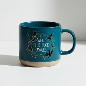 May include: Teal ceramic mug with a beige base and a large handle. The mug features a design of black dragons, a crescent moon, and the text "WELL THE FUCK AWARE" in white. The design is surrounded by gold stars and clouds.