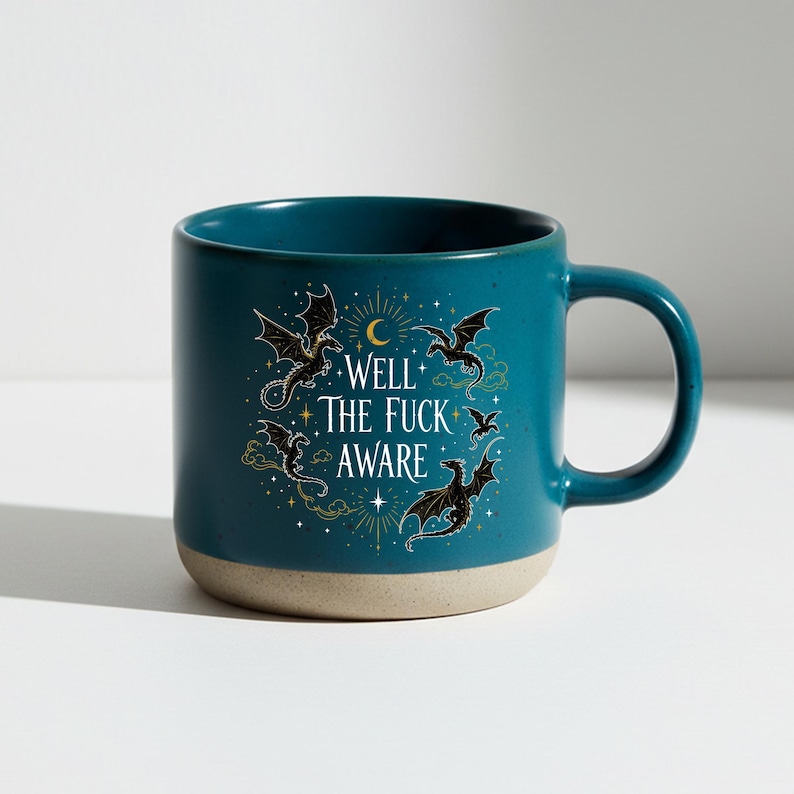 May include: Teal ceramic mug with a beige base and a large handle. The mug features a design of black dragons, a crescent moon, and the text "WELL THE FUCK AWARE" in white. The design is surrounded by gold stars and clouds.