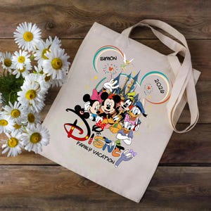 K&ouml;nnte beinhalten: Beige Tragetasche mit einem Disney-Familienurlaubsdesign. Das Design umfasst Micky und Minnie Maus, Donald Duck, Goofy und Daisy Duck. Der Text "Disney Family Vacation" befindet sich unter den Figuren. Das Jahr "2026" ist ebenfalls enthalten.