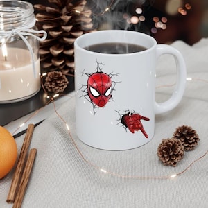 Puede incluir: Taza de cer&aacute;mica blanca con un dise&ntilde;o 3D de la m&aacute;scara y la mano de Spider-Man rompiendo una pared. La taza contiene un l&iacute;quido oscuro. El dise&ntilde;o es rojo y negro.