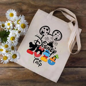 Puede incluir: Un bolso tote de color crema con un dise&ntilde;o negro de Disney que presenta las orejas de Mickey Mouse, fuegos artificiales y la palabra "Disney". Debajo, ilustraciones coloridas de Mickey, Minnie, Donald y Goofy. La palabra "Trip" tambi&eacute;n est&aacute; impresa.