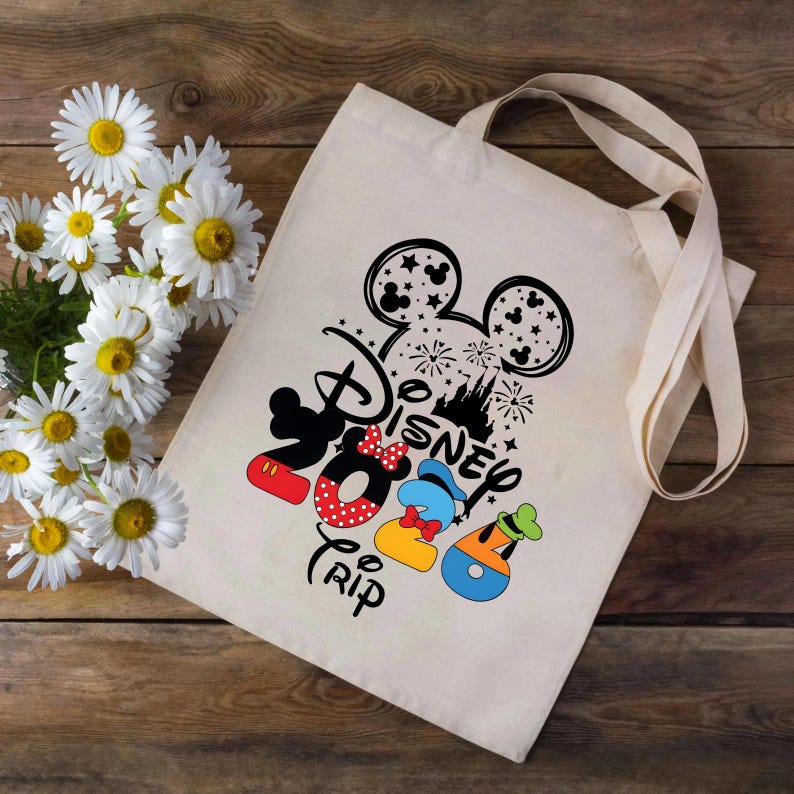 Puede incluir: Un bolso tote de color crema con un dise&ntilde;o negro de Disney que presenta las orejas de Mickey Mouse, fuegos artificiales y la palabra "Disney". Debajo, ilustraciones coloridas de Mickey, Minnie, Donald y Goofy. La palabra "Trip" tambi&eacute;n est&aacute; impresa.