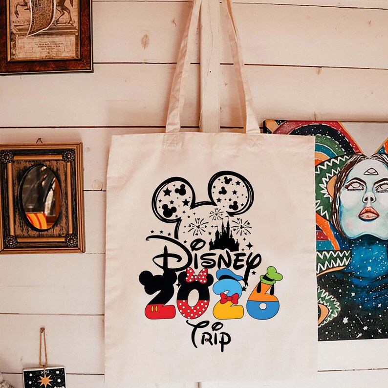 Puede incluir: Un bolso tote de color crema con una silueta negra de Mickey Mouse, fuegos artificiales y un dise&ntilde;o de castillo. Las palabras "Disney 2023 Trip" est&aacute;n impresas en negro, rojo, azul y amarillo. El bolso tiene dos asas color crema.