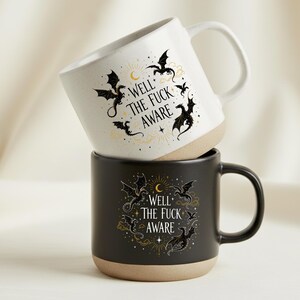 May include: Two ceramic mugs, one white and one black, stacked. Both mugs have a beige base and handle. The mugs feature a dragon design and the text "WELL THE FUCK AWARE". The mugs are designed for hot beverages.