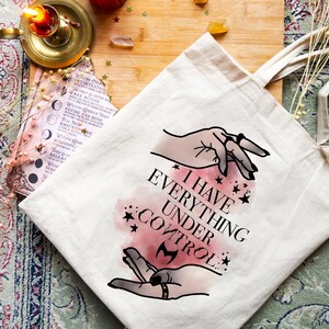 May include: A cream-coloured tote bag featuring the text "I HAVE EVERYTHING UNDER CONTROL" in black. The design includes illustrations of hands, stars, and a pink watercolour effect. The bag has long, beige handles.