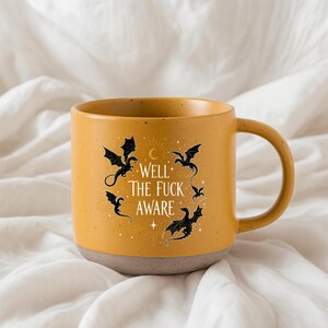 May include: Mustard yellow ceramic mug with a grey base. The mug features black dragon illustrations and the text "WELL THE FUCK AWARE" in a circular design. The mug has a large handle.