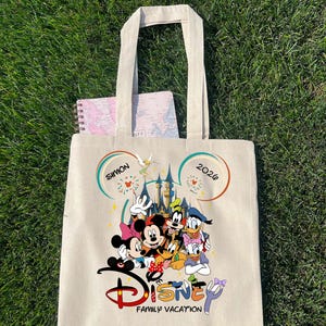 K&ouml;nnte beinhalten: Beige Canvas-Tragetasche mit farbenfrohem Disney-Familienurlaubsdesign. Das Design zeigt Micky und Minnie Maus, Donald Duck, Goofy und andere Charaktere vor einem Schloss. Der Text "Disney Family Vacation" ist darunter gedruckt.