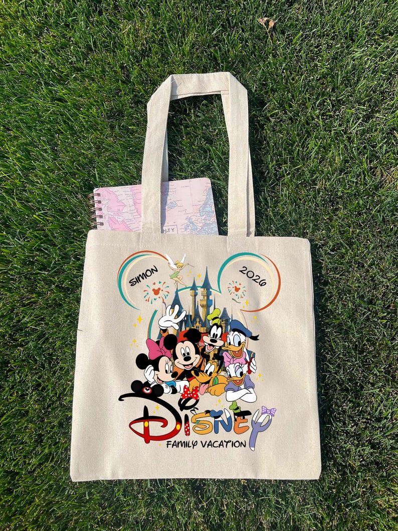 K&ouml;nnte beinhalten: Beige Canvas-Tragetasche mit farbenfrohem Disney-Familienurlaubsdesign. Das Design zeigt Micky und Minnie Maus, Donald Duck, Goofy und andere Charaktere vor einem Schloss. Der Text "Disney Family Vacation" ist darunter gedruckt.
