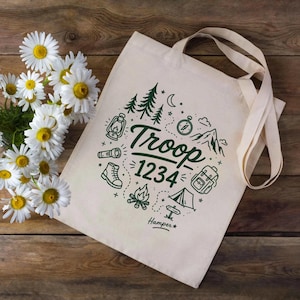 Op de afbeelding: Cr&egrave;mekleurige draagtas met een groen kampeerthema. Het ontwerp omvat bomen, een lantaarn, een kompas, een laars, een rugzak, een tent en de tekst "Troop 1234". De tas heeft lange handvatten en ligt op een houten oppervlak.