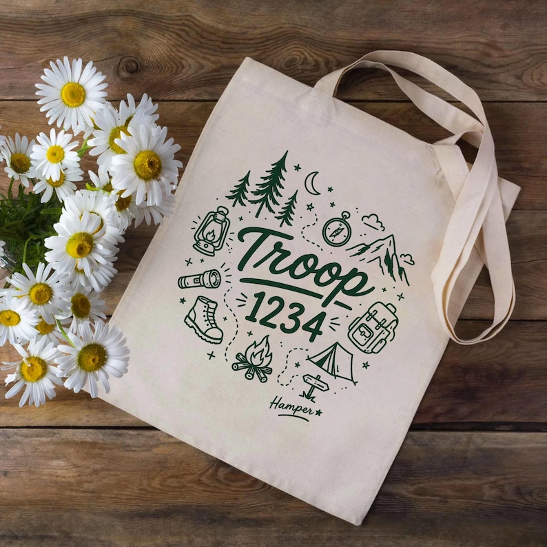 Op de afbeelding: Cr&egrave;mekleurige draagtas met een groen kampeerthema. Het ontwerp omvat bomen, een lantaarn, een kompas, een laars, een rugzak, een tent en de tekst "Troop 1234". De tas heeft lange handvatten en ligt op een houten oppervlak.