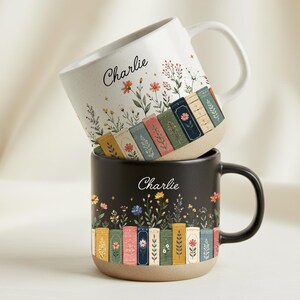Puede incluir: Dos tazas de cerámica apiladas. La taza superior es blanca con un diseño floral y de libros, y el nombre "Charlie" en cursiva. La taza inferior es negra con un diseño similar. Ambas tazas tienen una base beige y un asa curvada.