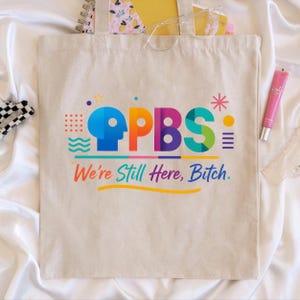 Könnte beinhalten: Eine beigefarbene Tragetasche mit dem farbenfrohen Text "PBS" und dem Satz "We're Still Here, Bitch." Das Design umfasst eine blaue Kopfsilhouette, orange, lila und grüne Buchstaben sowie verschiedene grafische Elemente. Die Tasche ist mit Notizbüchern und Brillen gefüllt.