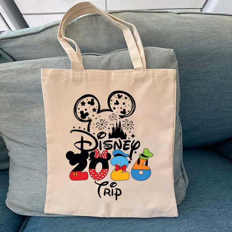 Puede incluir: Bolso tote de lona beige con un dise&ntilde;o de Disney. El dise&ntilde;o incluye orejas de Mickey Mouse, fuegos artificiales, una silueta de castillo y las palabras "Disney Trip 2023" con detalles de personajes. El bolso tiene dos asas beige.