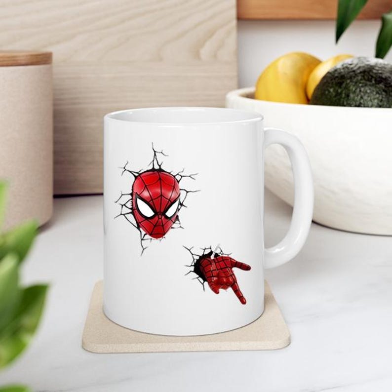 Puede incluir: Taza de cer&aacute;mica blanca con un dise&ntilde;o de Spider-Man rojo y negro. El dise&ntilde;o incluye la m&aacute;scara y la mano del personaje, como si rompieran la superficie de la taza. La taza est&aacute; sobre un posavasos beige.