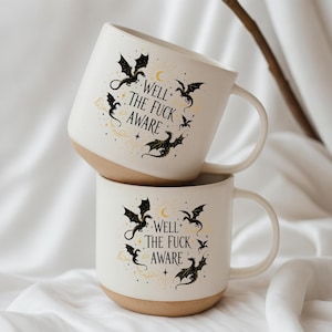 May include: Two stacked white ceramic mugs with a beige base. Each mug features a black dragon design with the text "Well the Fuck Aware" in a circular arrangement. The mugs have a curved handle.