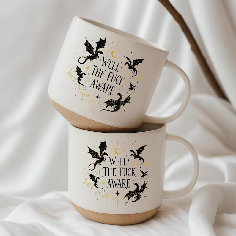 May include: Two stacked white ceramic mugs with a beige base. Each mug features a black dragon design with the text "Well the Fuck Aware" in a circular arrangement. The mugs have a curved handle.