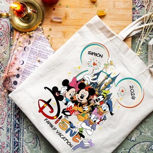 K&ouml;nnte beinhalten: Eine cremefarbene Tragetasche mit einem Disney-Familienurlaubsdesign. Das Design umfasst Micky und Minnie Maus, Donald und Daisy Duck und Goofy. Die Tasche tr&auml;gt den Text "Disney Family Vacation", den Namen "Simon" und das Jahr "2026".